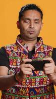 Vertical Happy Indian man entertained by videogames on smartphone, enjoying leisure time. Gamer enjoying game on phone, having fun defeating enemies, isolated over studio background, camera A video