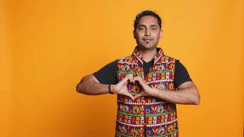 Portrait of smiling loving man doing heart symbol shape gesture with hands, showing kindness. Happy affectionate person doing tender love gesturing, isolated over studio background, camera B video