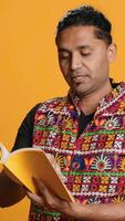Vertical Indian man reading book, having fun and enjoying characters and plot, isolated over studio background. Relaxed person browsing novel, enjoying leisure time, appreciating literature, camera A video