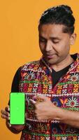 Vertical Influencer presenting green screen mobile phone, isolated over studio background. Indian person holding copy space chroma key smartphone used for advertising brands, camera A video