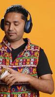 Vertical Joyous man listening music, having fun dancing, enjoying hot beverage. Upbeat person wearing headphones, doing funky movements, drinking coffee, studio background, camera A video