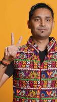 Vertical Portrait of upbeat man crossing fingers to make wish, hoping for good luck, isolated over studio background. Jolly indian person doing peace victory hand sign gesture, camera A video