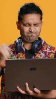 Vertical Indian man composing email, typing on laptop, frustrated with malfunctioning keyboard. Person sending mail online, struggling to use defective notebook, studio background, camera A video