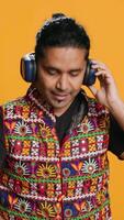 Vertical Indian man playing music, having fun moving on rhythm, pretending to use turntables. Upbeat DJ wearing headphones, producing beats, isolated over studio background, camera A video