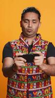 Vertical Happy Indian man entertained by videogames on smartphone, enjoying leisure time. Gamer enjoying game on phone, having fun defeating enemies, isolated over studio background, camera B video