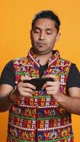 Vertical Cheerful Indian person in traditional clothing entertained by videogames on smartphone. Joyous player enjoying game on phone, having fun online, studio backdrop, camera B video