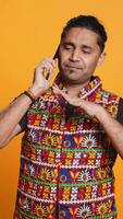 Vertical Happy man in traditional clothing chatting with best friend over telephone call. Upbeat person with colorful attire communicating with mate using phone, studio background, camera B video