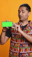 Vertical Content creator in traditional clothing recommending isolated screen phone, studio backdrop. Indian man reviewing copy space mockup smartphone device for online tech channel, camera B video