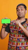 Vertical Smiling indian man presenting cellphone with green screen display, isolated over studio background. Person in traditional clothing creating promotion with blank copy space mockup phone, camera B video