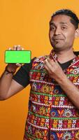 Vertical Content creator presenting green screen mobile phone, isolated over studio background. Indian person reviewing copy space mockup smartphone device for internet channel, camera B video