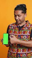 Vertical Influencer presenting green screen mobile phone, isolated over studio background. Indian person holding copy space chroma key smartphone used for advertising brands, camera B video