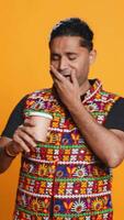 Vertical Tired indian man sipping fresh coffee from disposable paper cup early in the morning to wake up and be energized. Person drinking hot beverage from recycled takeaway cup, studio background, camera B video