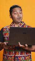 Vertical Indian man having entertaining conversation with friends during teleconference meeting using laptop, studio background. Happy person interacts with mates during online videocall on notebook, camera B video