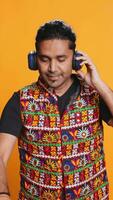 Vertical Indian man playing music, having fun moving on rhythm, pretending to use turntables. Upbeat DJ wearing headphones, producing beats, isolated over studio background, camera B video