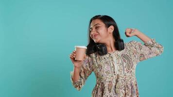 Joyful indian woman stretching and sipping fresh coffee from disposable paper cup after waking up. Radiant lively person drinking hot beverage early in the morning, studio background, camera A video