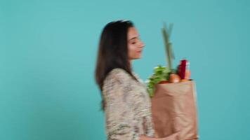 Woman walking with paper bag in hands filled with vegetables, living healthy lifestyle. Conscious living person holding groceries shopping bag, isolated over studio background video
