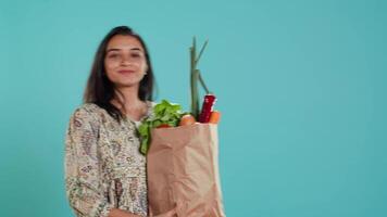 Happy indian woman walking with eco friendly paper bag in hands filled with vegetables, living healthy lifestyle. Portrait of smiling person holding bio groceries shopping bag, studio background video