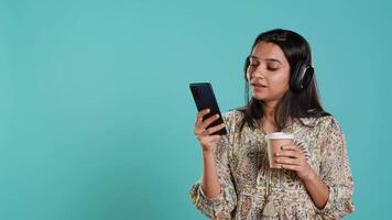 jubiloso mujer escuchando música, canto letra desde teléfono pantalla, disfrutando bebida. alegre persona vistiendo auriculares, escuchando canciones, cabeza saltando y participación café taza, estudio fondo, cámara un video