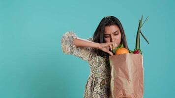 Indisch vrouw met ecologisch papier zak in handen op zoek Bij klok peper, leven gezond levensstijl. ecologie minnaar Holding boodschappen doen zak met biologisch produceren, studio achtergrond, camera een video