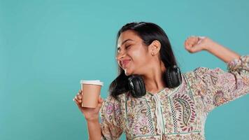 Joyful indian woman stretching and sipping fresh coffee from disposable paper cup after waking up. Radiant lively person drinking hot beverage early in the morning, studio background, camera B video