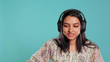 Happy woman playing electronic music and waving hand in air, pretending to use turntables. Cheerful DJ wearing headphones, producing beats, making songs, isolated over studio background, camera B video