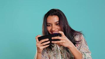 indio mujer jugando videojuegos en Teléfono móvil en paisaje modo, disfrutando ocio tiempo. jugador disfrutando juego en móvil teléfono, teniendo divertido, aislado terminado estudio fondo, cámara si video