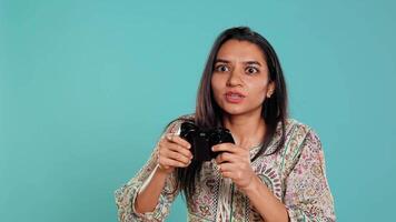 Jolly gamer celebrating after winning game on gaming console, studio background. Delighted Indian woman bragging after being victorious in videogame, defeating all enemies using gamepad, camera B video