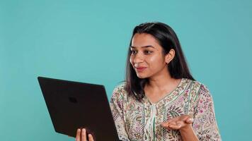 Upbeat woman saluting coworkers, having friendly conversations during teleconference meeting using laptop, studio background. Person discussing during online videocall on notebook device, camera B video