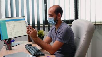 Entrepreneur texing on smartphone wearing mask while working in office room with new normal. Freelancer sitting in modern workspace chatting talking writing using mobile phone with internet technology video