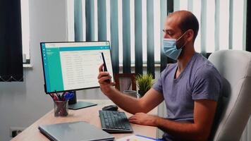 hombre con proteccion máscara utilizando teléfono inteligente leva en oficina habitación. persona de libre dedicación trabajando en nuevo normal lugar de trabajo chateando teniendo virtual conferencia, reunión con remoto colegas utilizando Internet video