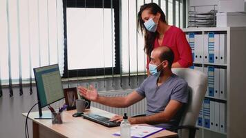 företag team arbetssätt bär skydd mask i ny vanligt kontor rum. anställda i modern företags- företag arbetsyta skriver på dator tangentbord och pekande på skrivbordet analyserar klienter lista video