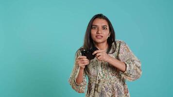 Jolly gamer celebrating after winning game on gaming console, studio background. Delighted Indian woman bragging after being victorious in videogame, defeating all enemies using gamepad, camera A video