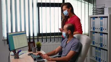 Mann mit Schutz Maske zeigen Kunden aufführen auf Computer Sitzung im Büro Zimmer. Angestellte Arbeiten im Neu normal Büro Arbeitsplatz Tippen auf Tastatur zeigen beim Desktop Analysieren Termine video