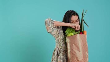 Indian woman with paper bag with vegetables and fruits testing quality, looking at orange, studio background. Vegetarian verifying groceries are ripe after buying them from zero waste shop, camera A video