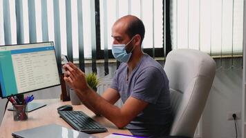 Man sitting in office room on desk typing on phone wearing protection mask. Freelancer working in new normal workspace chatting talking writing using mobile phone with internet technology video