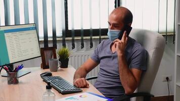Worker with face mask talking on phone in new modern office room. Freelancer working in modern workplace chatting writing speaking on smartphone with remotely colleagues sitting in front of computer video