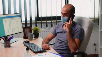 lavoratore con viso maschera parlando su Telefono nel nuovo moderno ufficio camera. libero professionista Lavorando nel moderno posto di lavoro chat scrittura A proposito di su smartphone con remoto colleghi seduta nel davanti di computer video