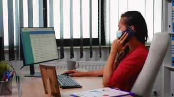 Entrepreneur with protection face mask talking on phone in front of computer. Hispanic freelancer working in new normal workplace speaking on smartphone with remotely colleagues looking at desktop video