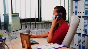 Woman with protection mask making appointments using mobile phone. Freelancer working in new normal workplace speaking on smartphone with remotely colleagues looking at desktop analyzing clients list video