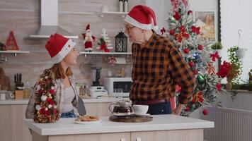 Grandchild with grandfather bringing xmas wrapper present gift with ribbon on it celebrating christmastime during winter holiday. Happy family enjoying christmas in decorated kitchen video