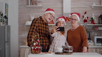 contento abuelos en pie a mesa en Navidad decorado cocina tomando selfie utilizando inteligente durante invierno estación. familia vistiendo Papa Noel sombrero haciendo gracioso expresiones disfrutando Navidad fiesta video