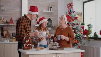 Grandparents enjoying spending time with grandchild celebrating christmastime eating baked chocolate delicious cookies discussing in xmas decorated kitchen. Family enjoying christmas holiday video