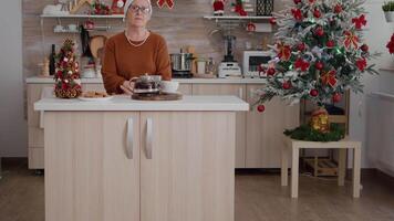Portrait of old woman wearing xmas hat celebrating christmas season in decorated kitchen enjoying winter traditional holiday. On table standing cup of coffee with baked delicious cookies video