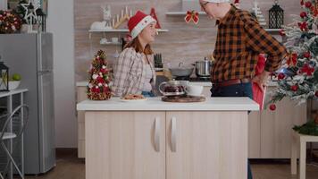 Happy family bringing wrapper christmas present gift with ribbon on it in xmas decorated kitchen celebrating christmas holiday. Grandfather with grandchild wearing santa hat enjoying winter season video