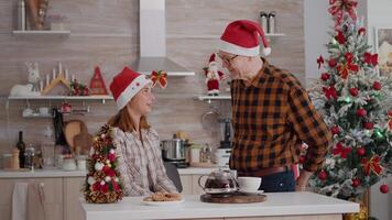 Grandfather surprising granddaughter with wrapper present gift in xmas decorated kitchen enjoying christmastime together. Happy family wearing santa hat celebrating christmas holiday video