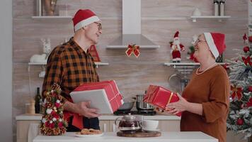 nonna condivisione involucro regalo presente con nastro su esso con nonno in piedi a tavolo nel natale decorato cucina durante inverno vacanza. contento famiglia godendo Natale stagione video