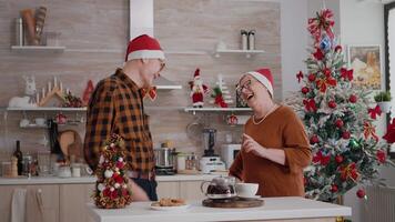 Happy grandparents couple family sharing wrapper gift with ribbon on it enjoying christmastime. Family celebrating winter holiday season surprinsing with present in decorated kitchen video