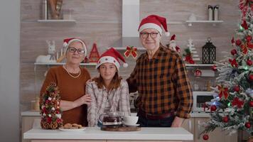 retrato do feliz família vestindo santa chapéu olhando para dentro Câmera em pé às mesa dentro natal decorado cozinha. avós desfrutando inverno estação gastos Natal feriado com neto video