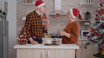 neta trazendo embrulho presente presente surpresa para avós a comemorar época de Natal dentro decorado cozinha. feliz família vestindo santa chapéu desfrutando inverno feriado durante Natal video