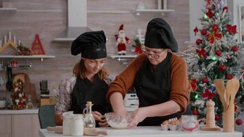 Grandmother showing to grandchild how to prepare traditional cookies dessert enjoying christmastime together. Child wearing apron while making homemade biscuits dough in home kitchen video